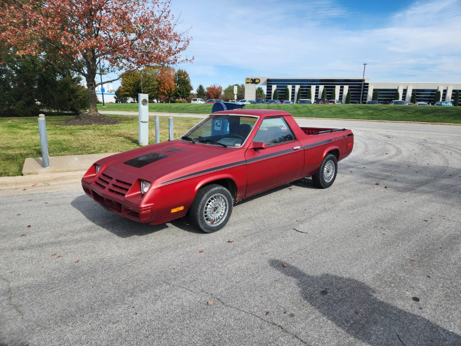 A red coupe utility vehicle is parked on a paved area near a grassy lawn, with trees and a modern office building in the background under a partly cloudy sky.
