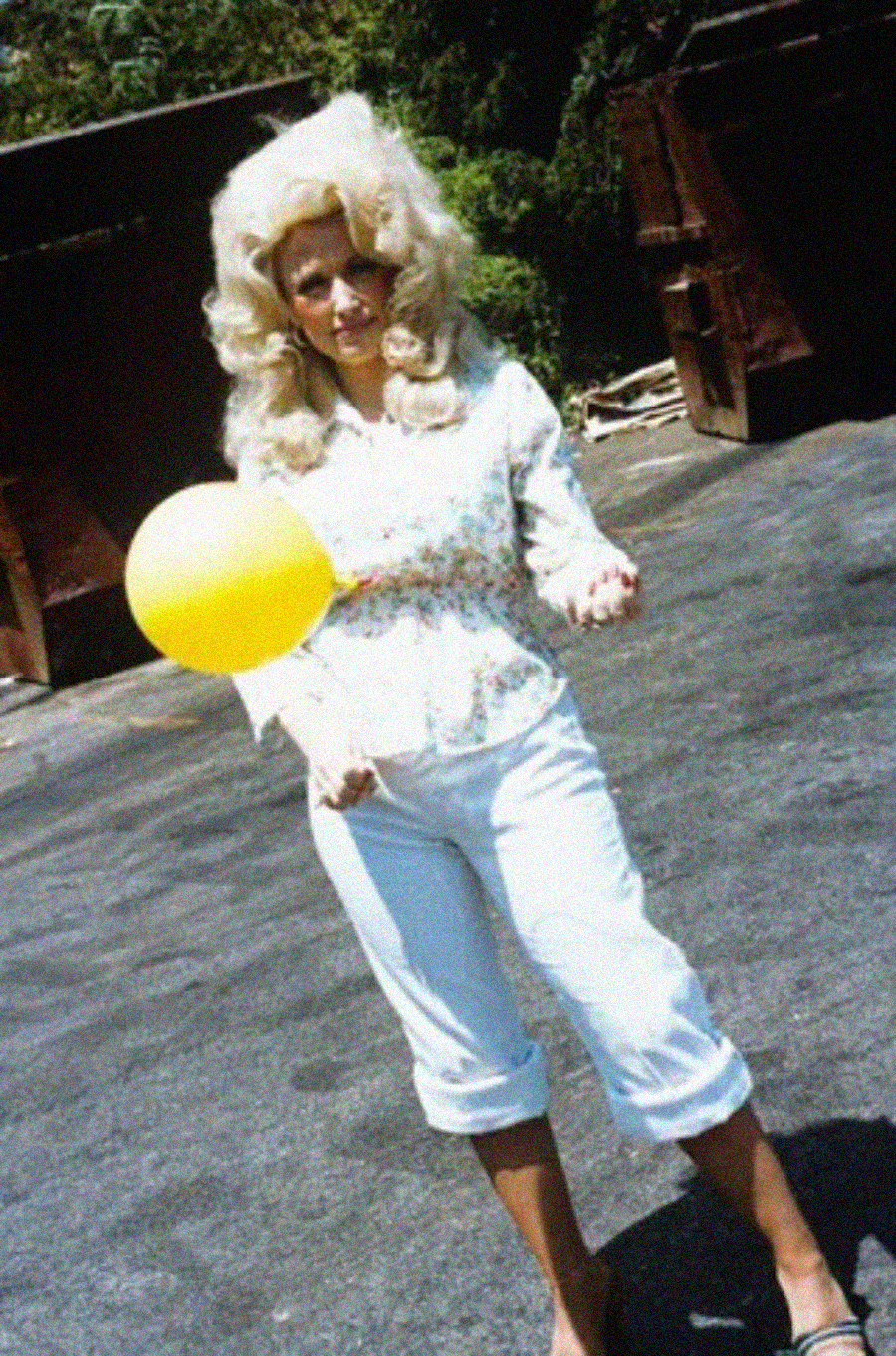 A woman with voluminous blonde hair, wearing a light floral blouse and white capri pants, stands outdoors on pavement holding a yellow balloon. Large brown dumpsters and greenery are visible in the background.