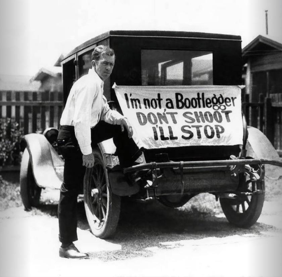 A man stands next to an old car with a sign on the back that reads, "I'm not a Bootlegger. DON’T SHOOT I’LL STOP." The scene appears to be from the early 20th century.