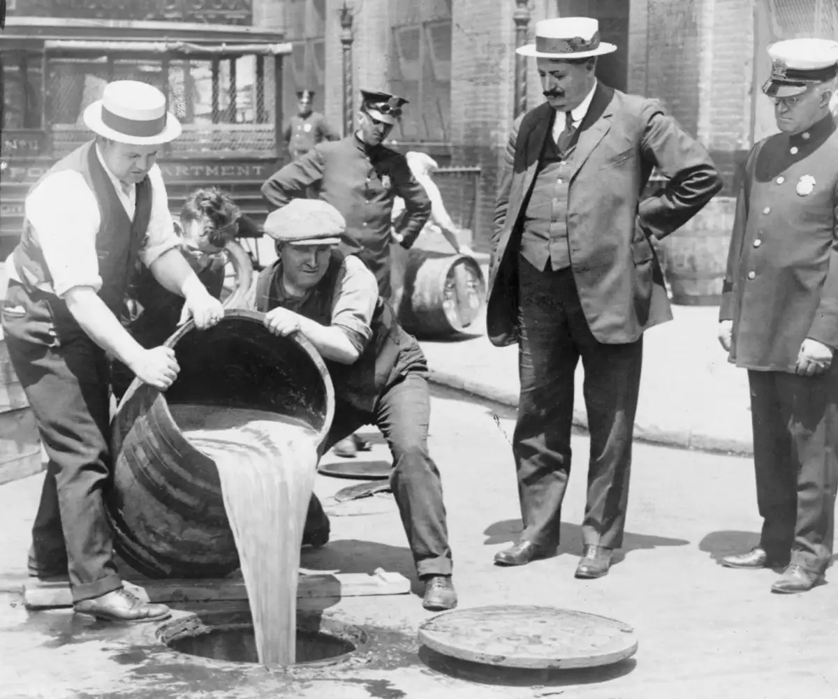 Four men, including police officers and officials, watch as two men pour liquid from a large barrel into a street drain during Prohibition, possibly destroying illegal alcohol. The scene takes place outdoors in an urban setting.