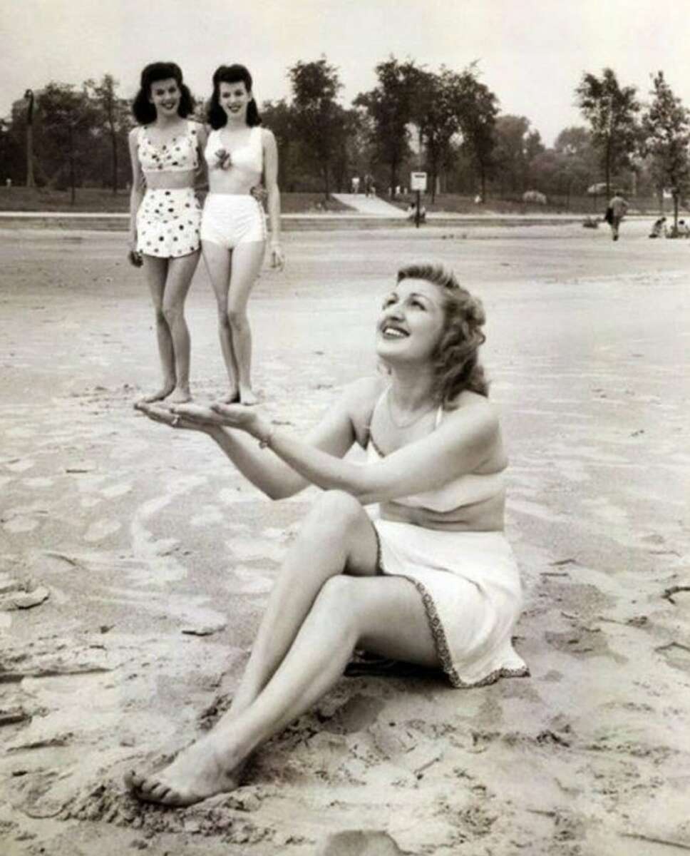 A woman sits on the beach smiling, holding out her hands as if supporting two distant women who appear tiny by perspective illusion. All three wear vintage swimsuits; trees and sand are visible in the background.