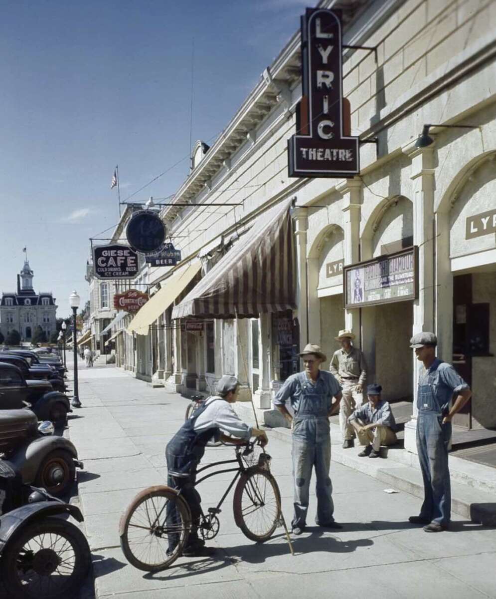 A vintage street scene shows four men in overalls talking outside the Lyric Theatre, with one leaning on a bicycle. Old cars are parked along the sidewalk near shops and a café under striped awnings.