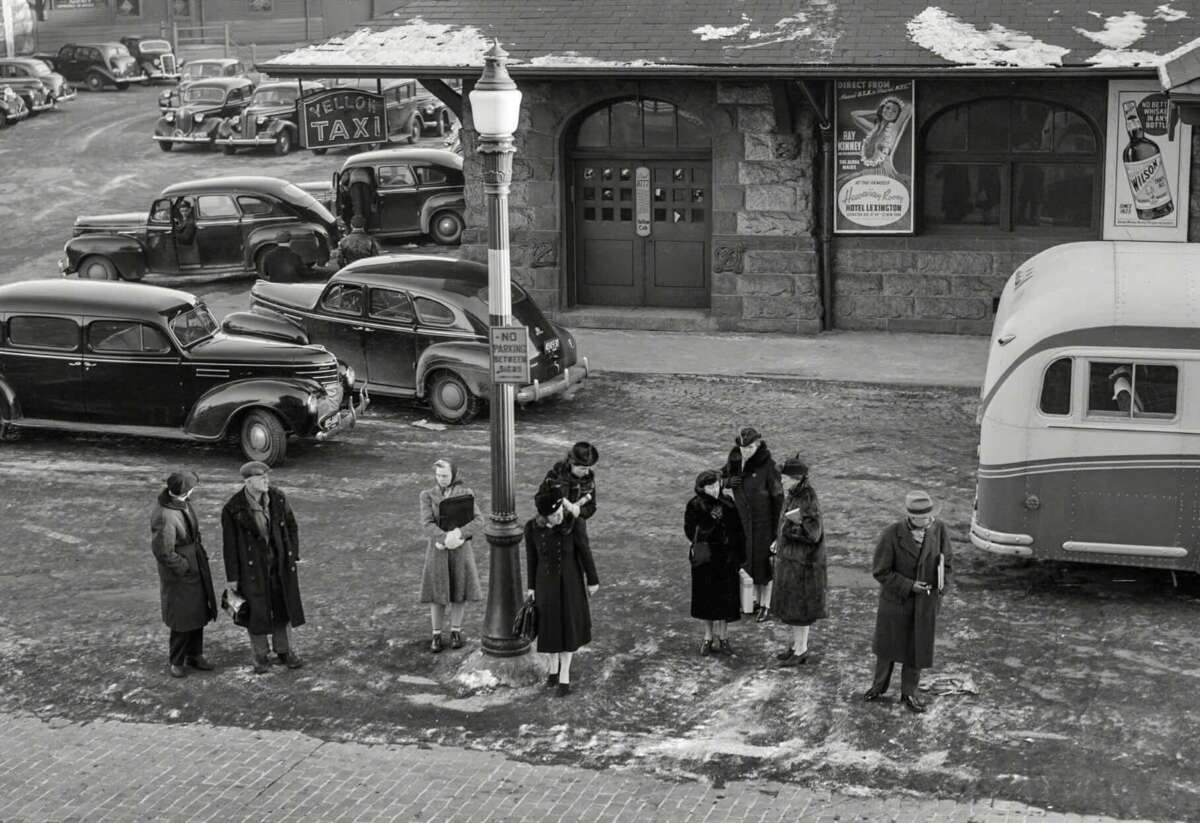 Black and white photo of people in winter coats waiting at a bus stop near a streetlamp, with vintage cars and a taxi stand in the background. Snow patches are visible on the ground and rooftops.