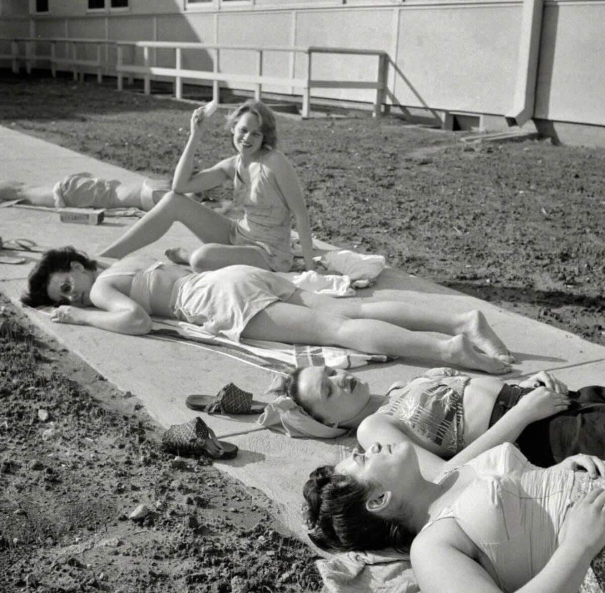 Four women in swimsuits sunbathe on towels on a concrete path next to a building, while another woman sits up and smiles at the camera. Sandals and personal items are scattered nearby.