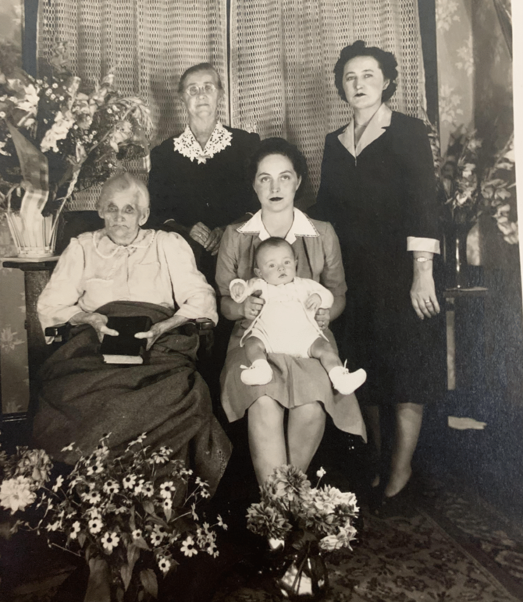Five women from different generations, including an elderly woman, two middle-aged women, a young woman holding a baby, and a toddler, pose indoors surrounded by flower arrangements. The photo is in black and white.