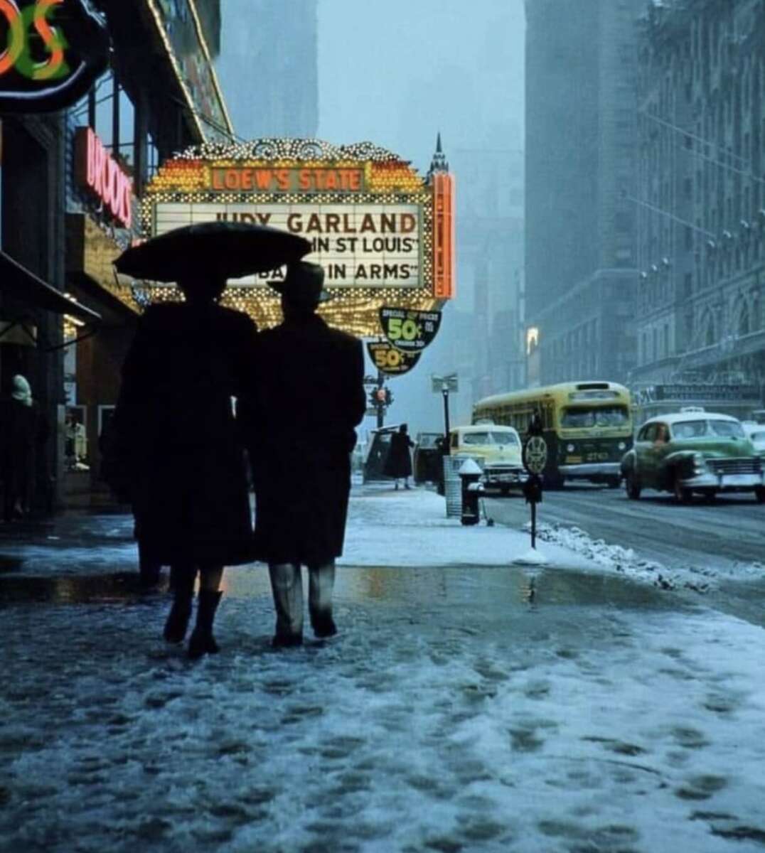 Two people walk under umbrellas on a snowy city sidewalk at dusk, passing a brightly lit theater marquee advertising Judy Garland in "Meet Me in St. Louis." Vintage cars line the wet street.