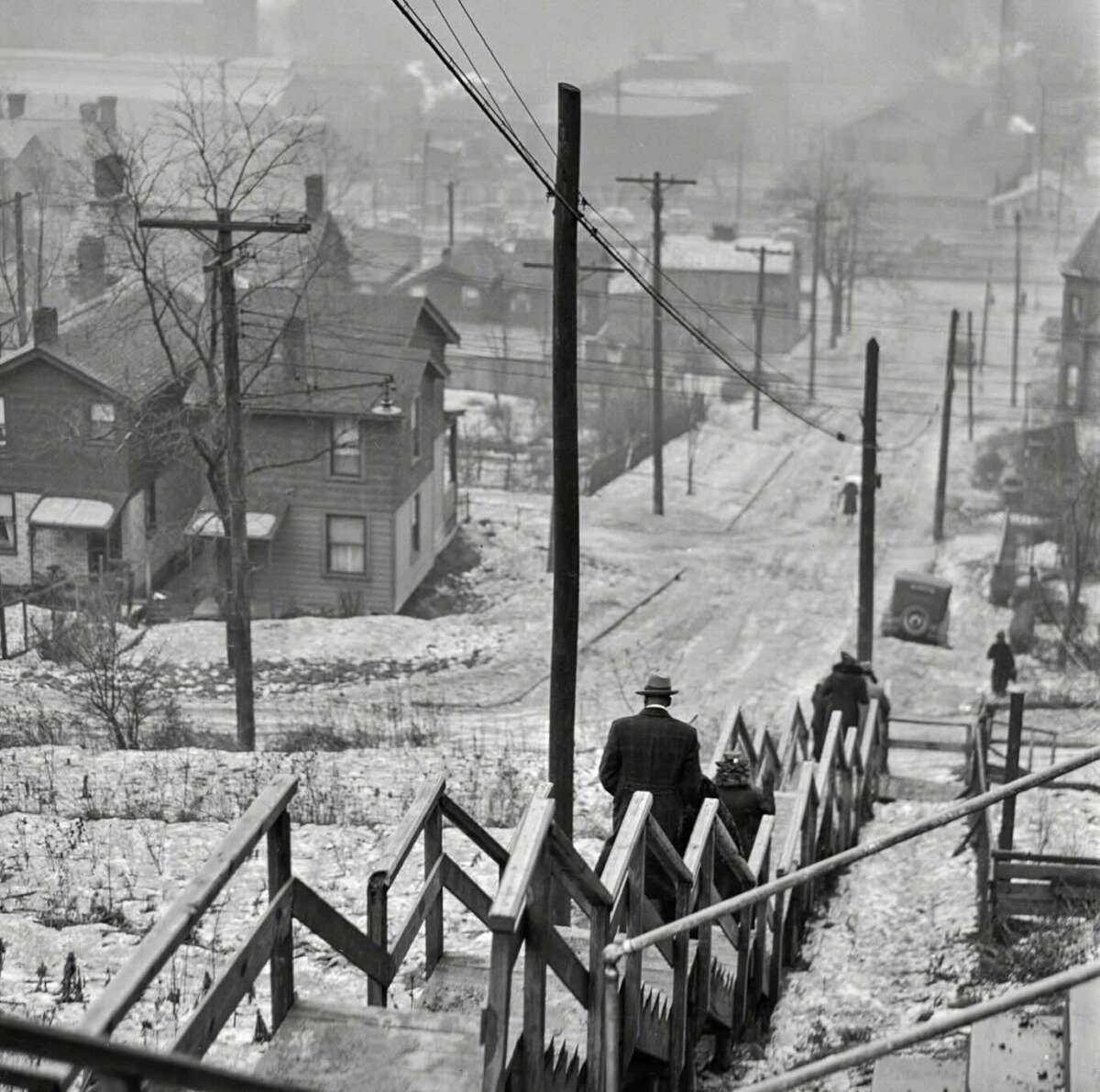 A man and a child walk down a steep, wooden staircase toward a snow-covered street lined with houses and utility poles on a foggy winter day. Crooked rooftops and distant figures are visible in the misty background.