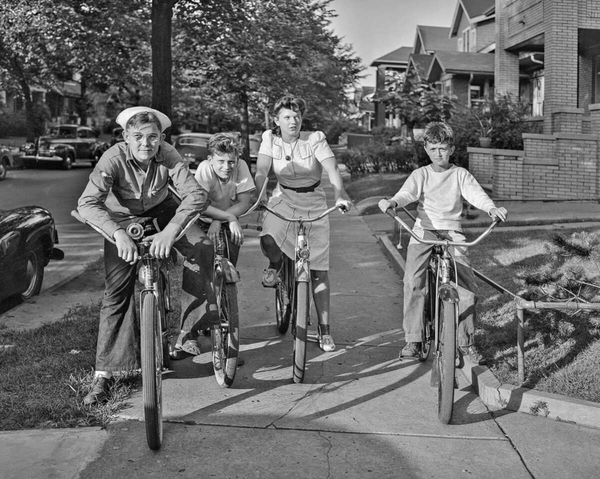 Four children, three boys and one girl, are riding bicycles on a sidewalk in a residential neighborhood with trees and parked cars, likely in the 1940s or 1950s. They look toward the camera, smiling and relaxed.