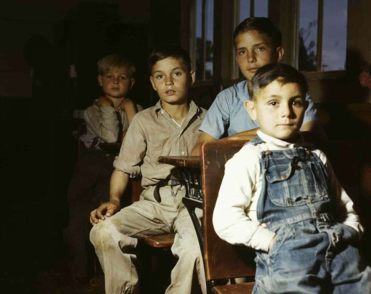Four boys sit and stand in a dimly lit classroom, wearing worn clothes and overalls. Two boys sit at a wooden desk while two others stand behind them, all looking toward the camera with neutral expressions.