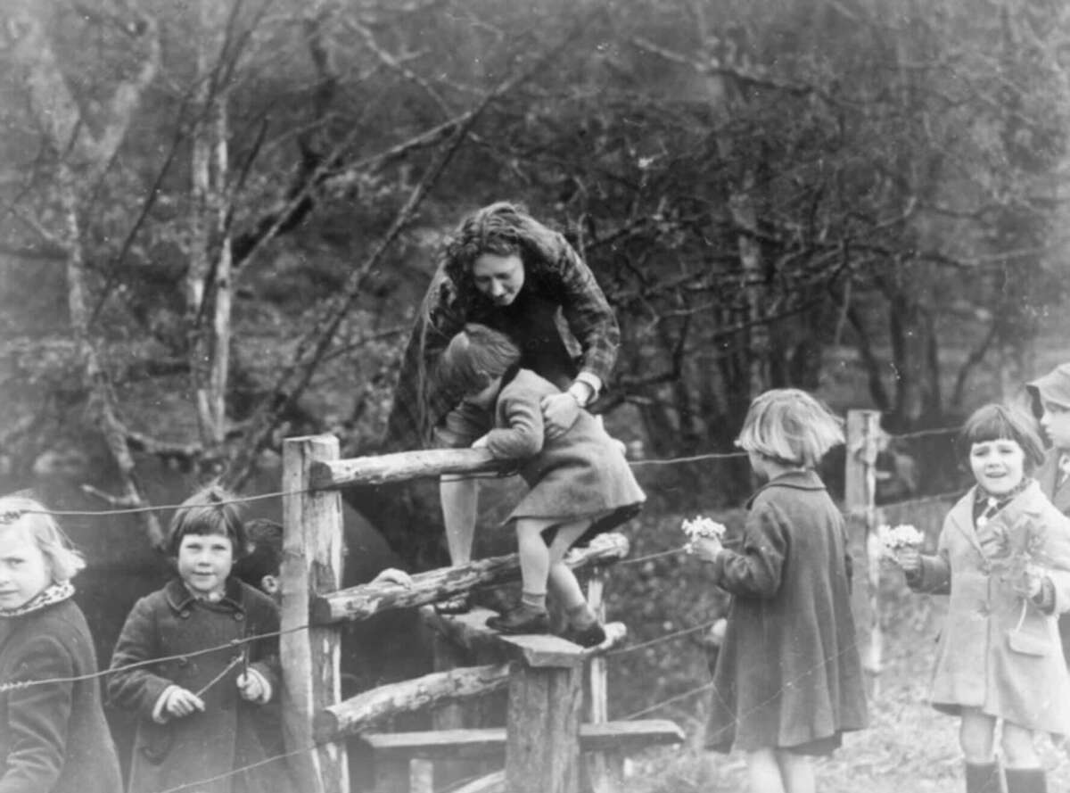 A woman helps a child climb over a wooden fence in a rural area, while several children holding flowers stand nearby, all dressed in coats. The background shows trees and foliage. The image is in black and white.