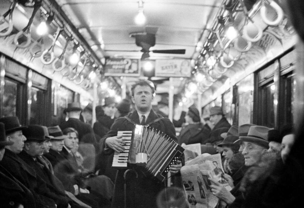 A man plays an accordion while standing in the aisle of a crowded vintage subway car filled with passengers, some seated and some standing, many wearing hats and coats.
