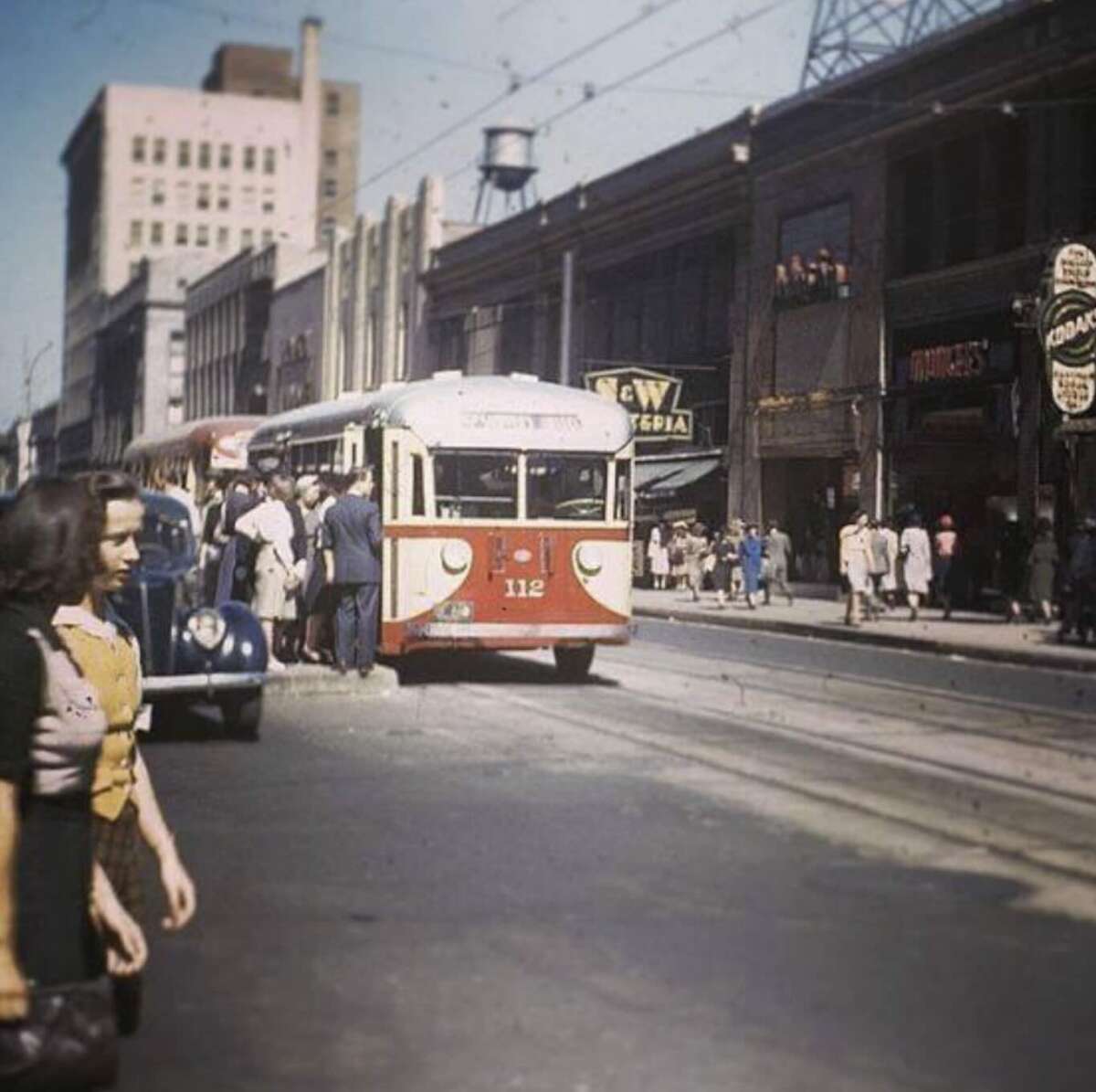 A vintage streetcar numbered 112 travels down a busy city street lined with shops and pedestrians, while a woman walks in the foreground and cars are parked along the curb.