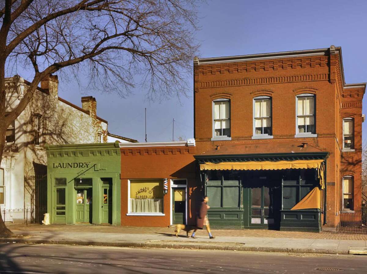 A person walks a dog past a small green building labeled "Laundry" and a larger red brick building with yellow awnings on a quiet street in sunlight, with bare trees casting shadows.