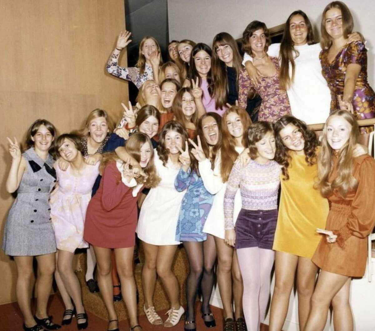 A group of young women in colorful 1960s or 1970s dresses smile and make peace signs while posing together on a staircase, showing excitement and joy.