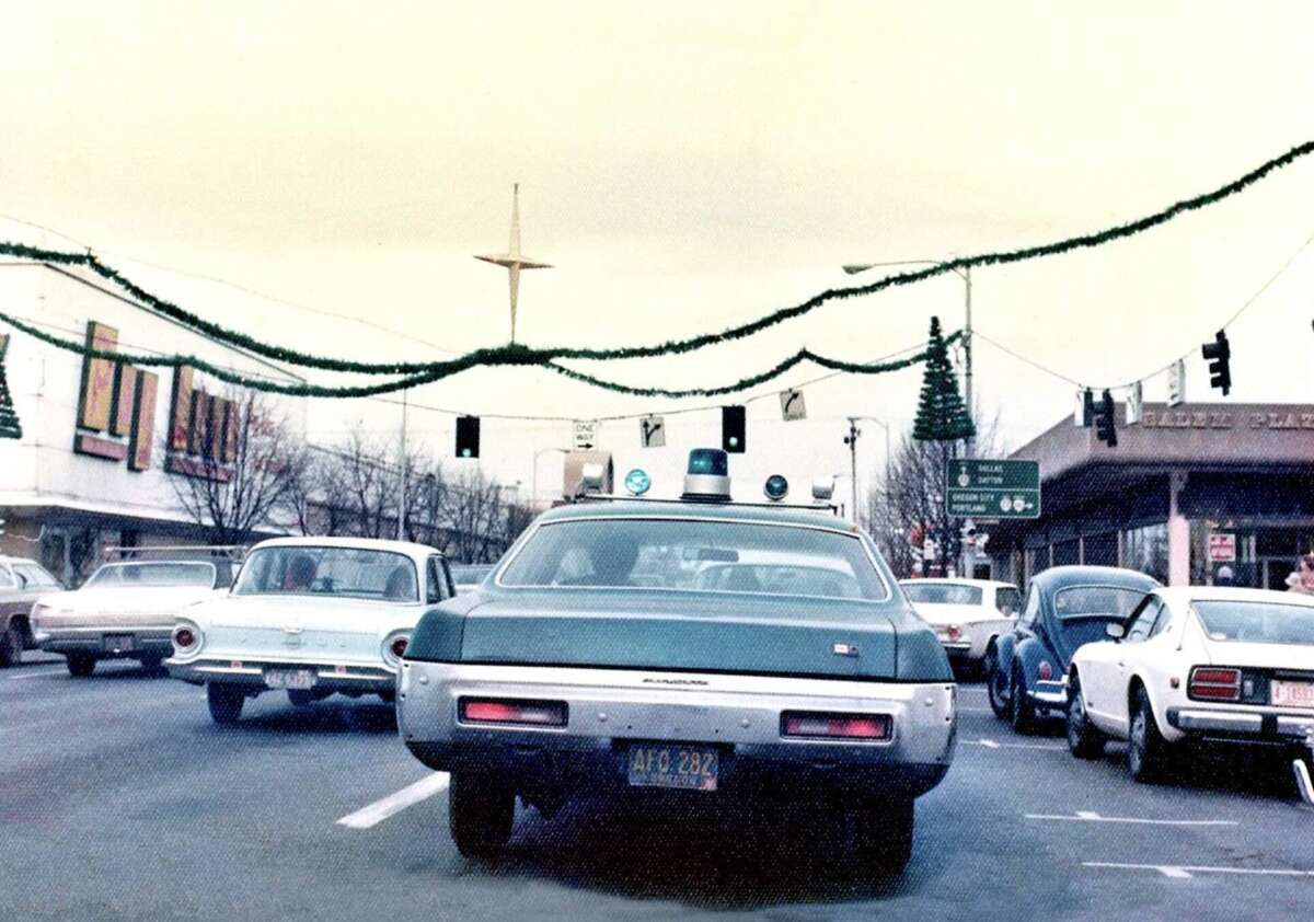 A vintage police car with roof lights drives down a city street decorated with holiday garlands and tree ornaments. Several classic cars are present and storefronts are visible under an overcast sky.