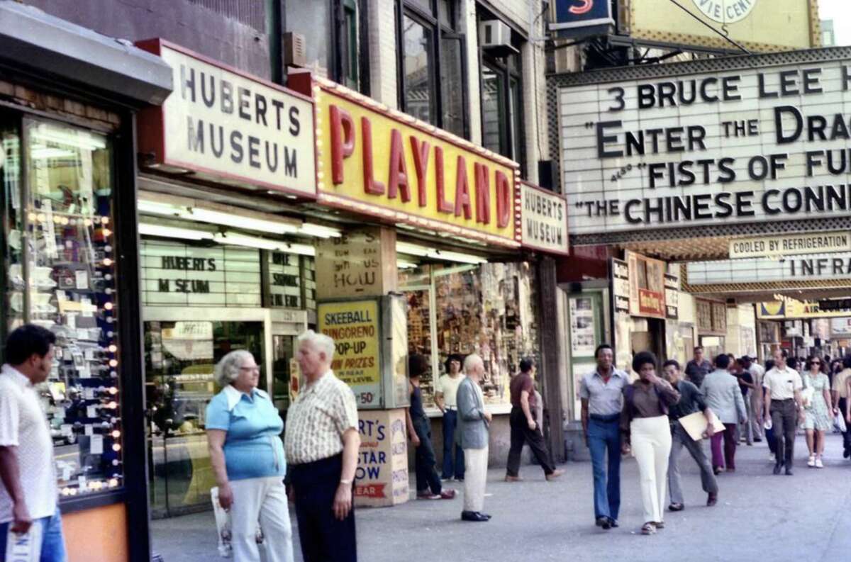 A busy city street scene from the 1970s with people walking past shops including Hubert’s Museum and Playland, and a theater marquee advertising Bruce Lee’s movies.