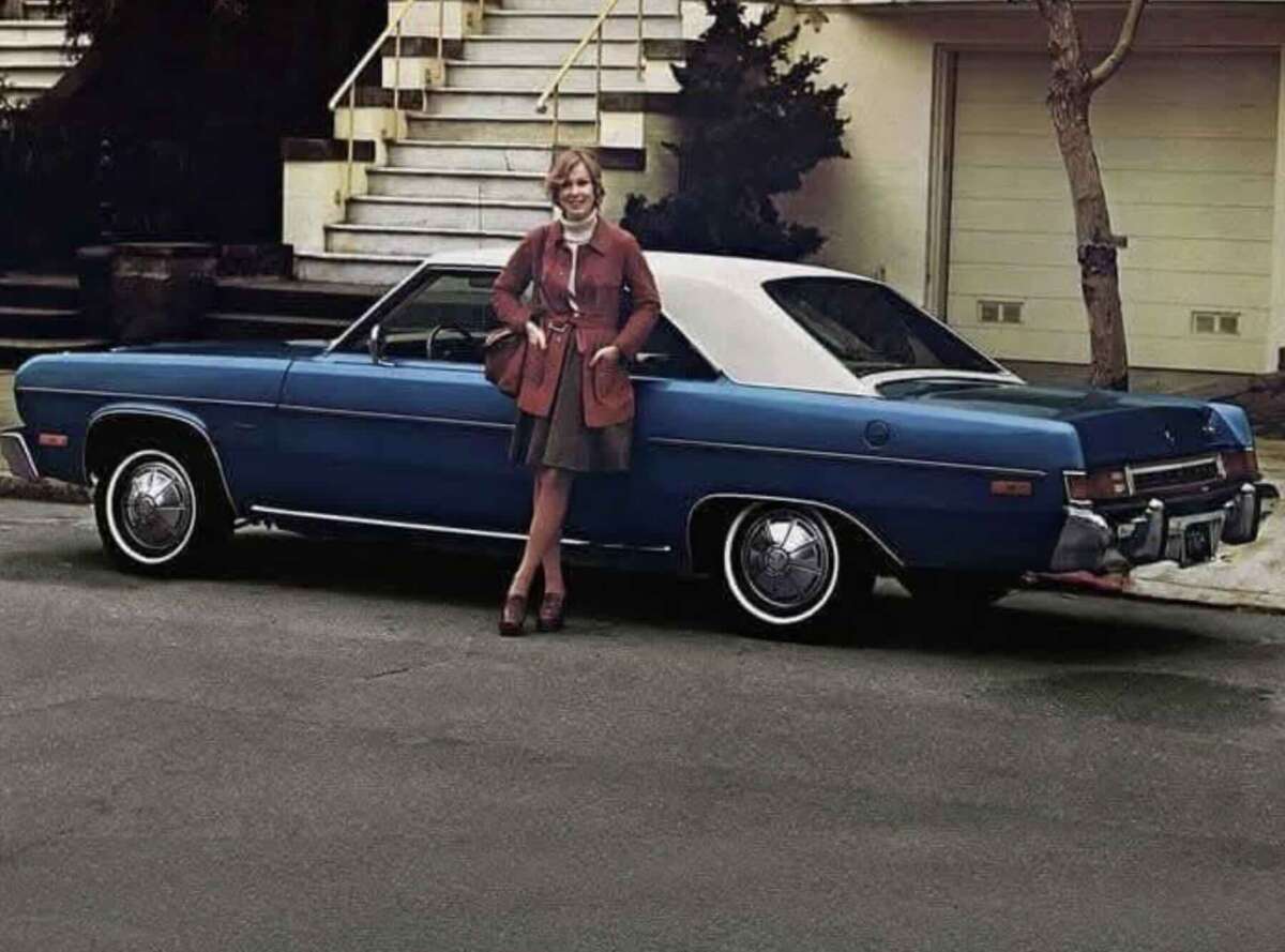 A woman in a red jacket and skirt stands smiling beside a classic blue and white two-door car parked on a suburban street near stairs and a garage.