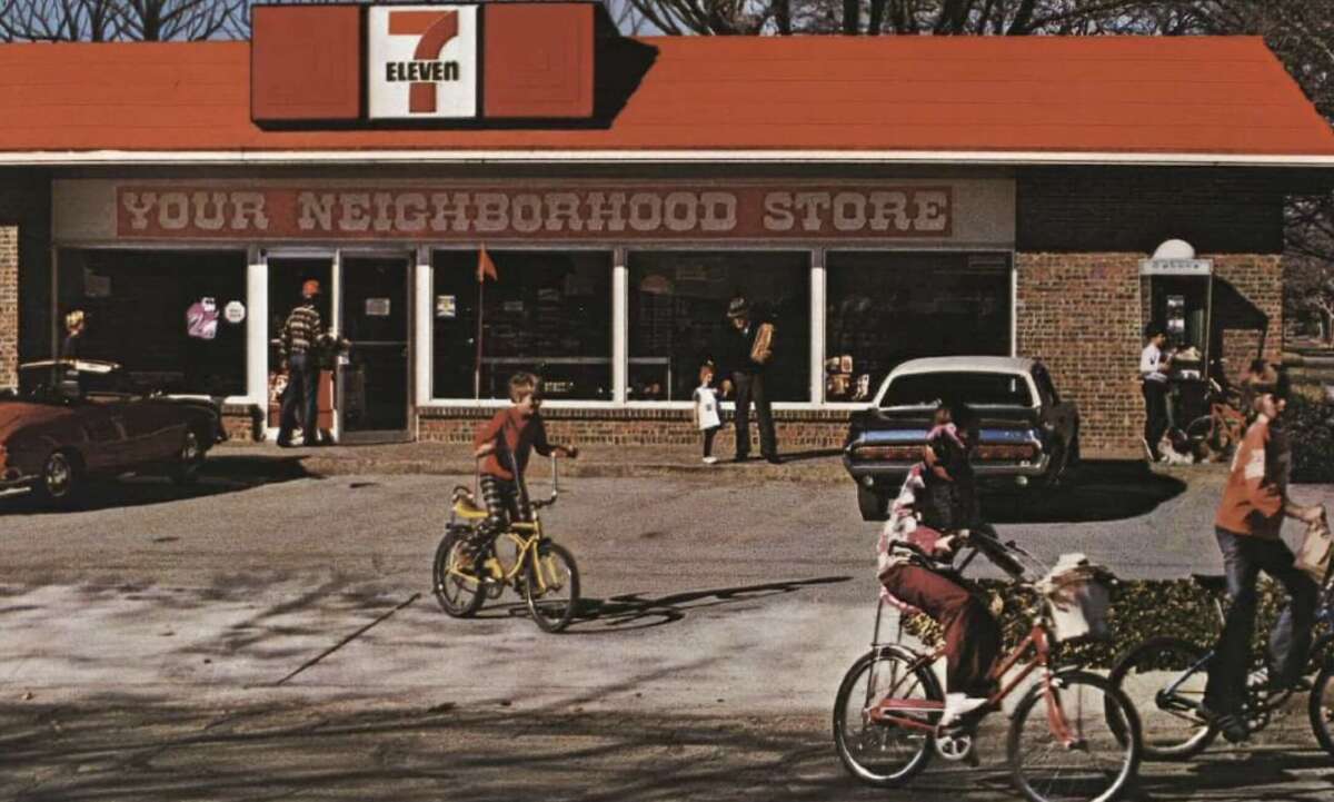 Children ride bicycles in front of a vintage 7-Eleven store with a red sign that reads "YOUR NEIGHBORHOOD STORE." Several cars are parked, and people walk near the entrance on a sunny day.