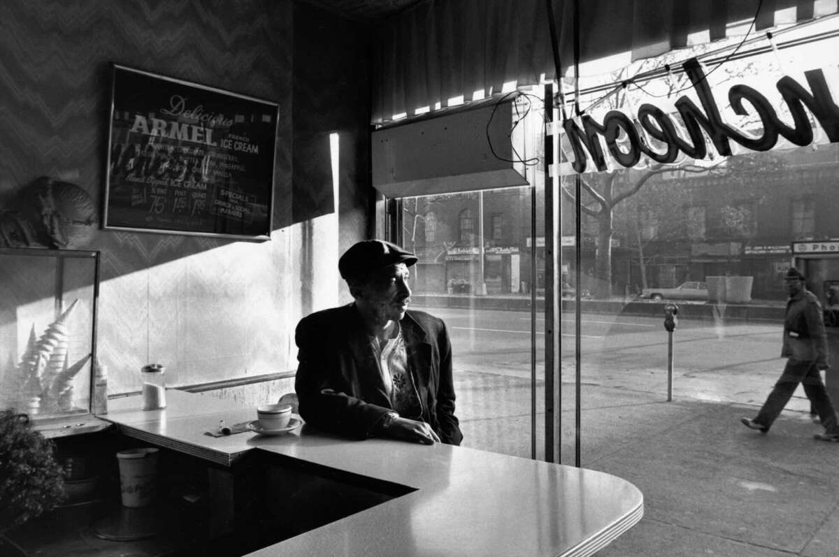 An older man wearing a cap sits alone at a counter in a diner with sunlight streaming through large windows. A cup and saucer are on the counter, and a pedestrian walks outside on the sidewalk.