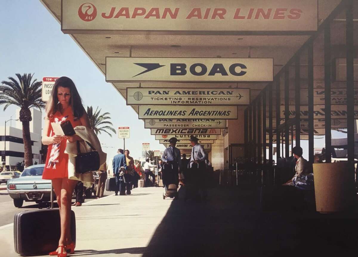 A woman in a red dress stands with luggage outside an airport terminal, reading a document. Signs for Japan Air Lines, BOAC, Pan American, and other airlines are visible above her. Palm trees and people are in the background.