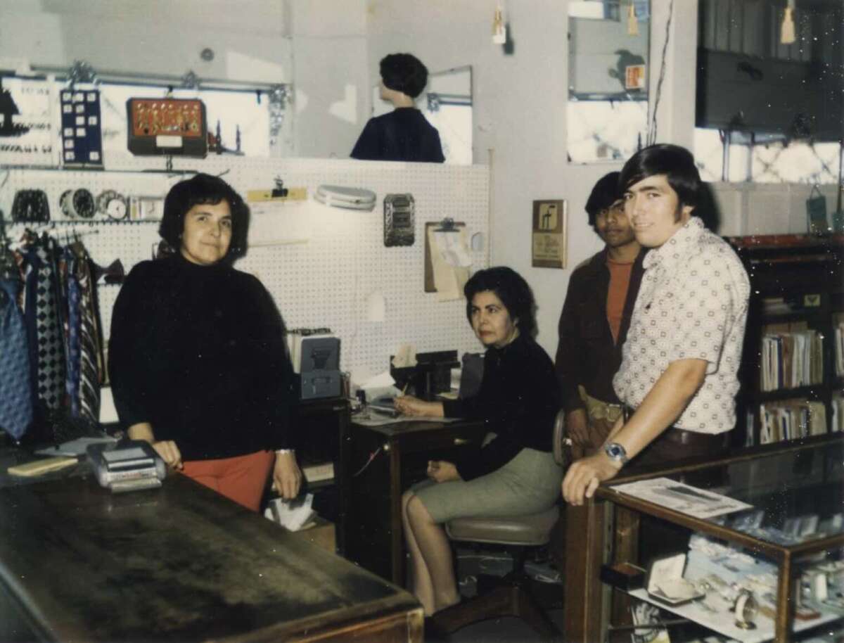 Four people pose inside a vintage shop, with clothing, books, and office equipment visible. Two women stand by the counter, one sits at a desk, and two men stand nearby. The atmosphere is casual and retro.