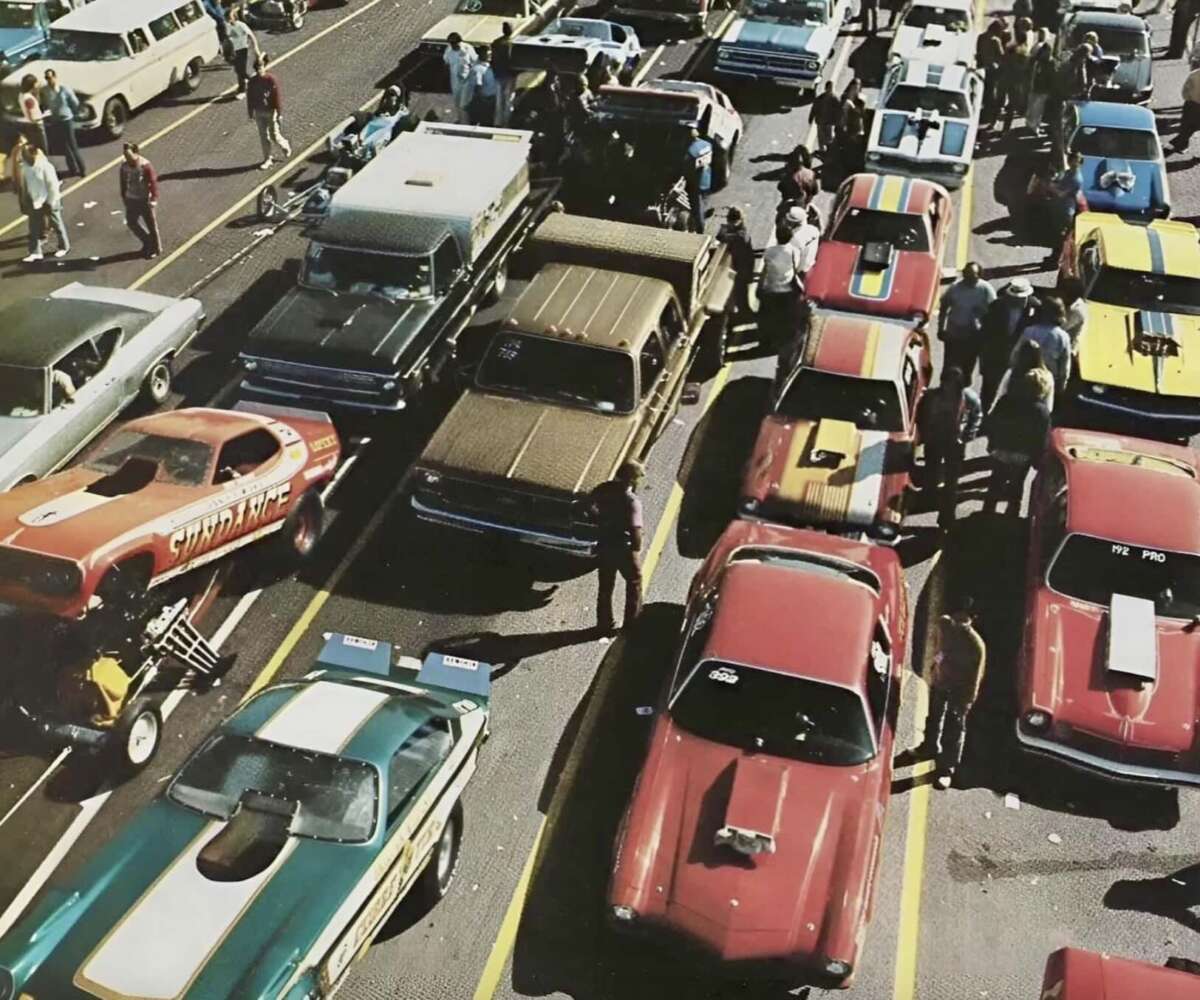 A crowded parking area at a car show or race event features rows of colorful classic cars and hot rods, with people walking and gathering around the vehicles on a sunny day.