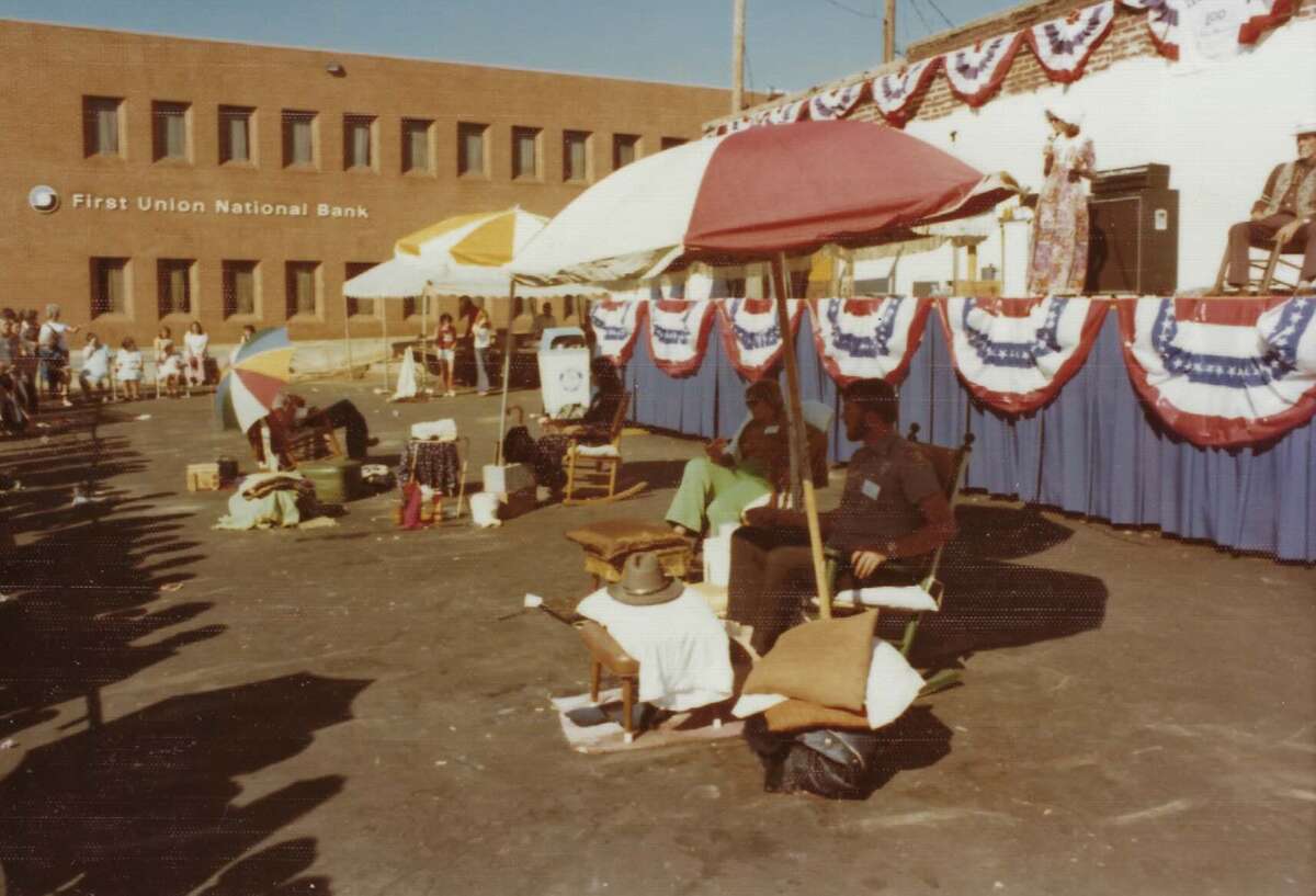 People sit under colorful umbrellas in front of a stage decorated with red, white, and blue bunting. A crowd looks on near a brick building labeled "First Union National Bank.
