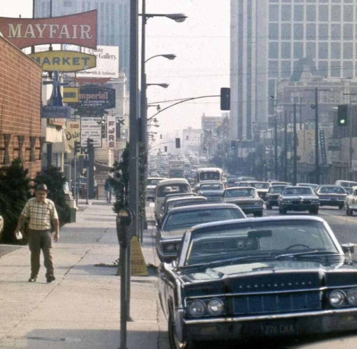 A busy city street from the 1970s, lined with parked cars and vintage signs. A man in a plaid shirt and hat walks on the sidewalk as traffic fills the road, with tall office buildings in the background.