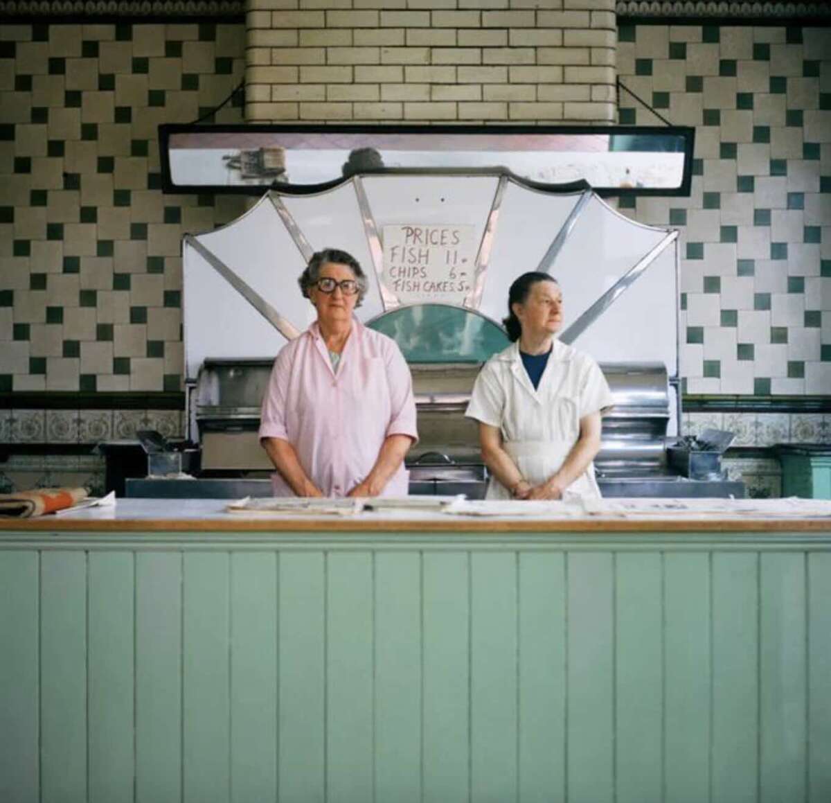 Two people in white uniforms stand behind the counter of a vintage-style fish and chip shop, with a sign listing prices for fish, chips, and fishcakes on the wall behind them.