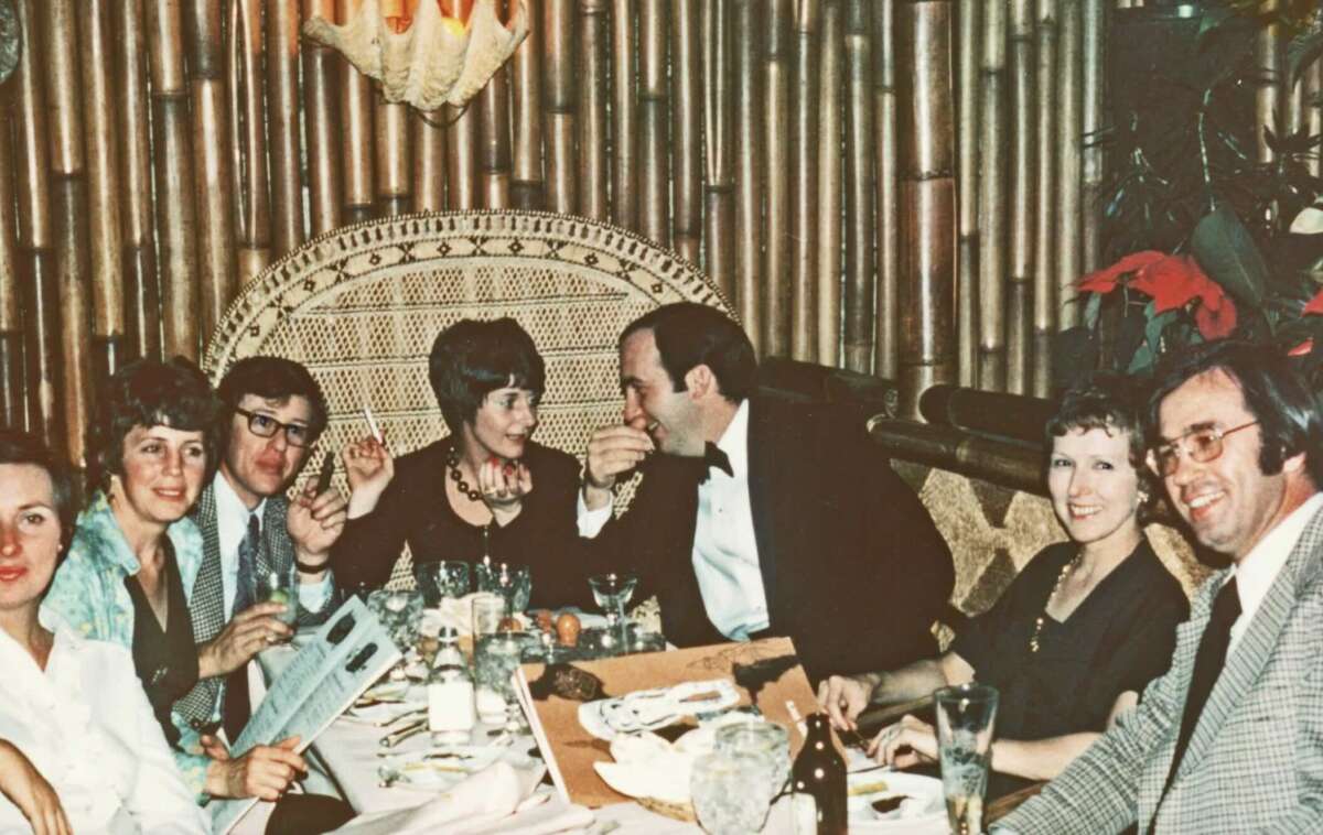 A group of seven adults sit around a table in a dimly lit restaurant, dressed in formal attire, smiling and talking, with bamboo decor and an ornate chair in the background.