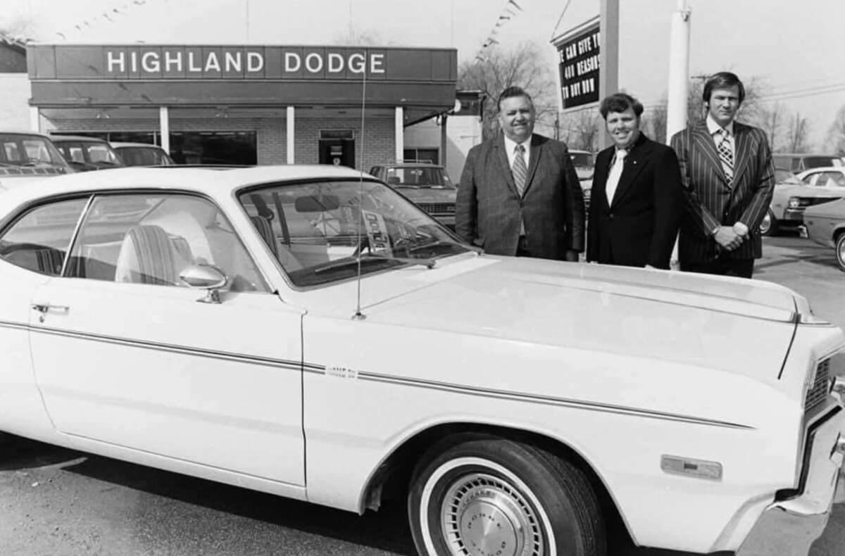 Three people in formal attire stand beside a classic white car in front of a dealership with a sign that reads "Highland Dodge." Other cars are visible in the background.