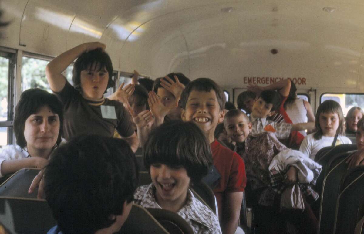 Children sit and laugh on a school bus, some making silly faces and hand gestures. The bus is full, and sunlight comes through the windows. The mood appears playful and joyful.