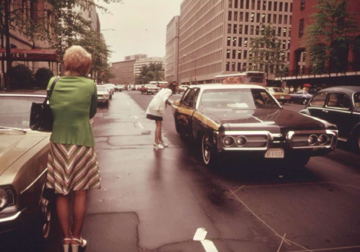A woman in a green jacket stands on a city street as another woman leans into the window of a parked car. The street is lined with cars and tall buildings, and the pavement appears wet from rain.