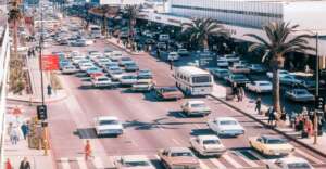 A busy city street from the mid-20th century with numerous vintage cars, a large white bus, palm trees, pedestrians, and storefronts along the sidewalk under bright daylight.