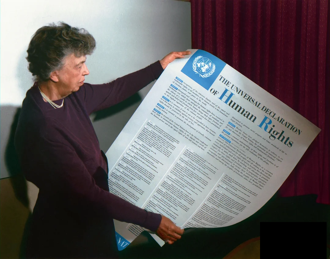 A woman stands holding a large printed copy of the Universal Declaration of Human Rights, reviewing the document. The text and the United Nations emblem are visible on the poster.