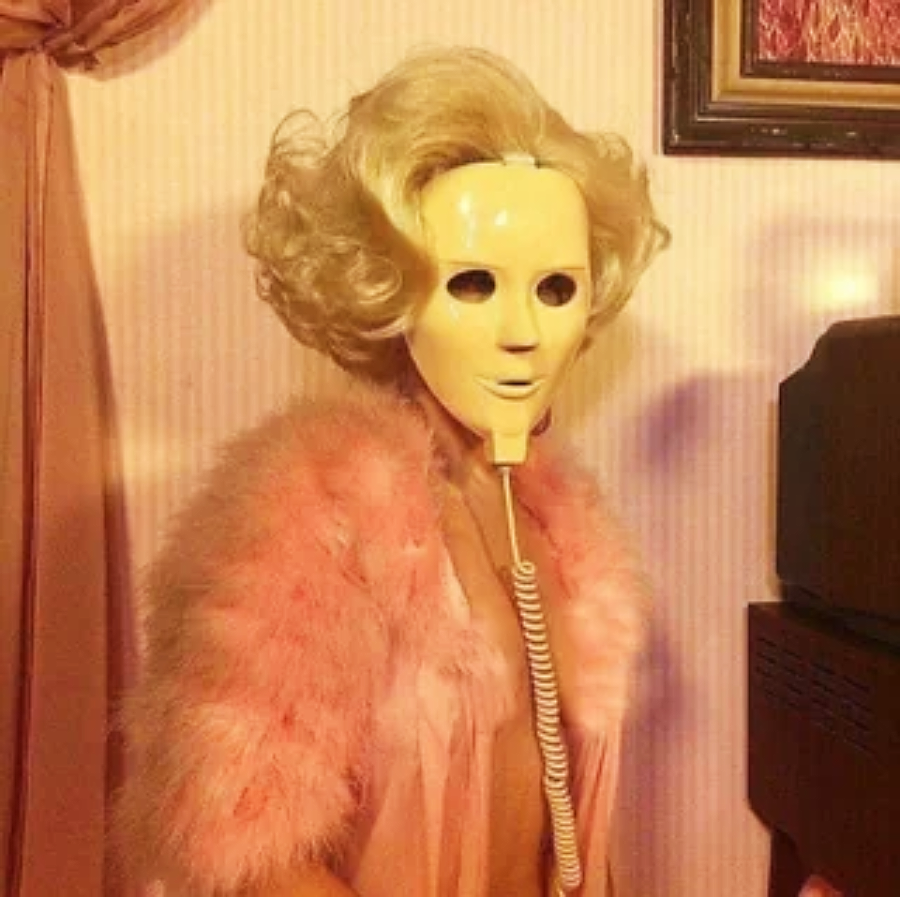 A person in a pink feathered robe wears a vintage, expressionless plastic mask with a tube attached to the mouth. The background includes striped wallpaper and a framed picture.