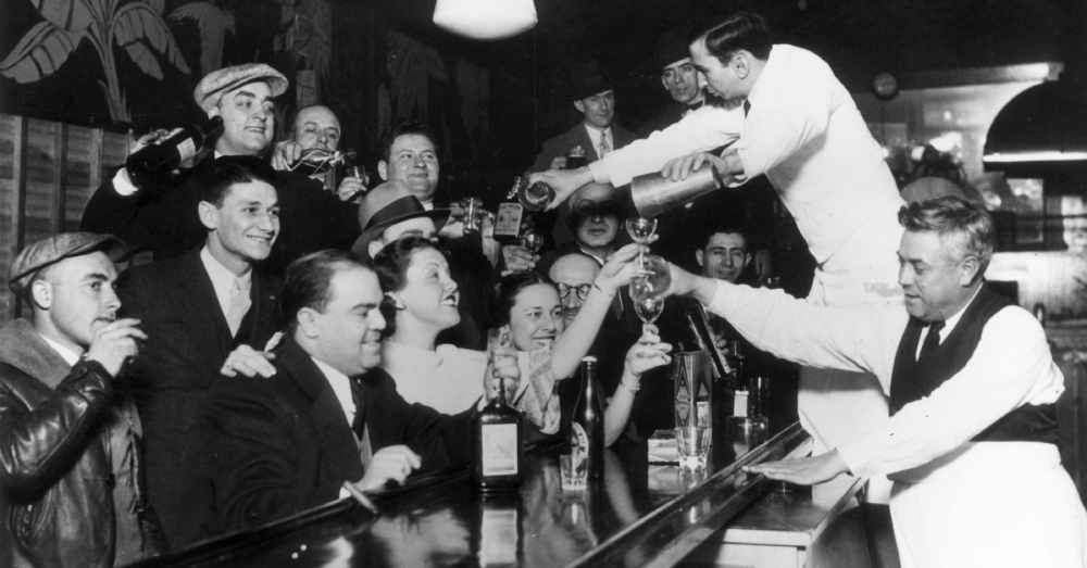 Black-and-white photo of a lively bar scene with men and women smiling, some raising drinks, as bartenders in white uniforms pour liquor into glasses behind the counter. The atmosphere is festive and crowded.