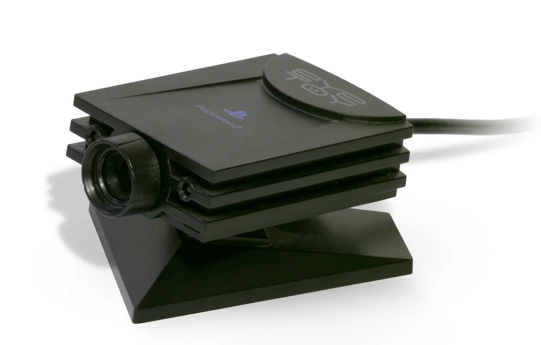 A black PlayStation 2 EyeToy camera with a lens on the front, USB cable attached, and PlayStation logo visible, resting on its stand against a white background.