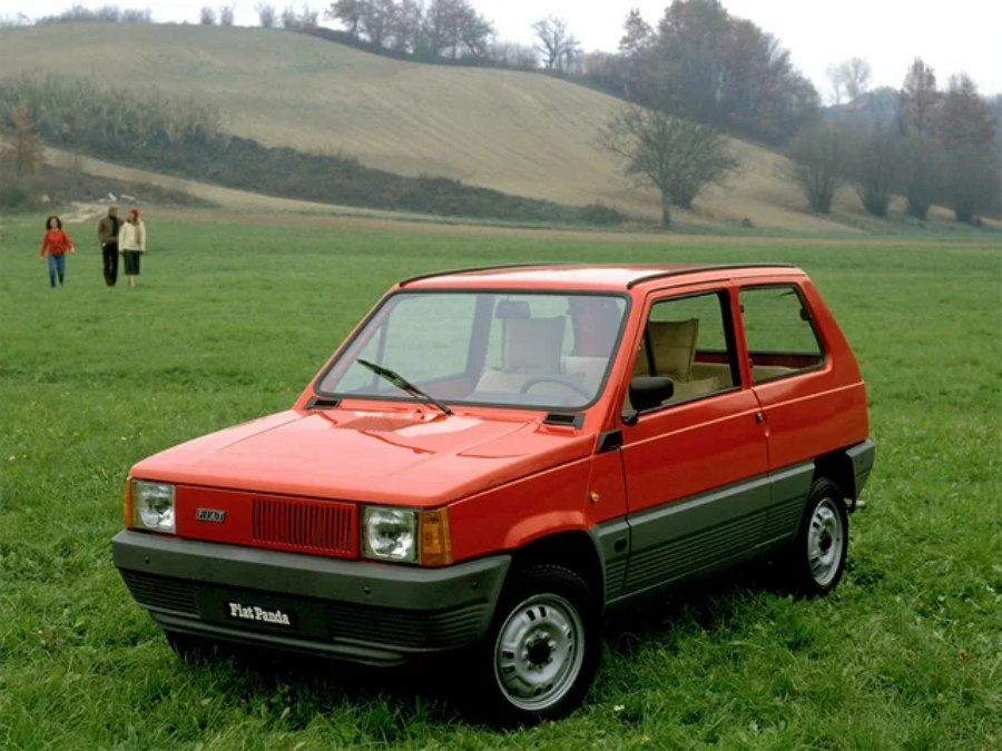 A red Fiat Panda hatchback is parked on a grassy field with trees and rolling hills in the background. Three people are walking in the distance.