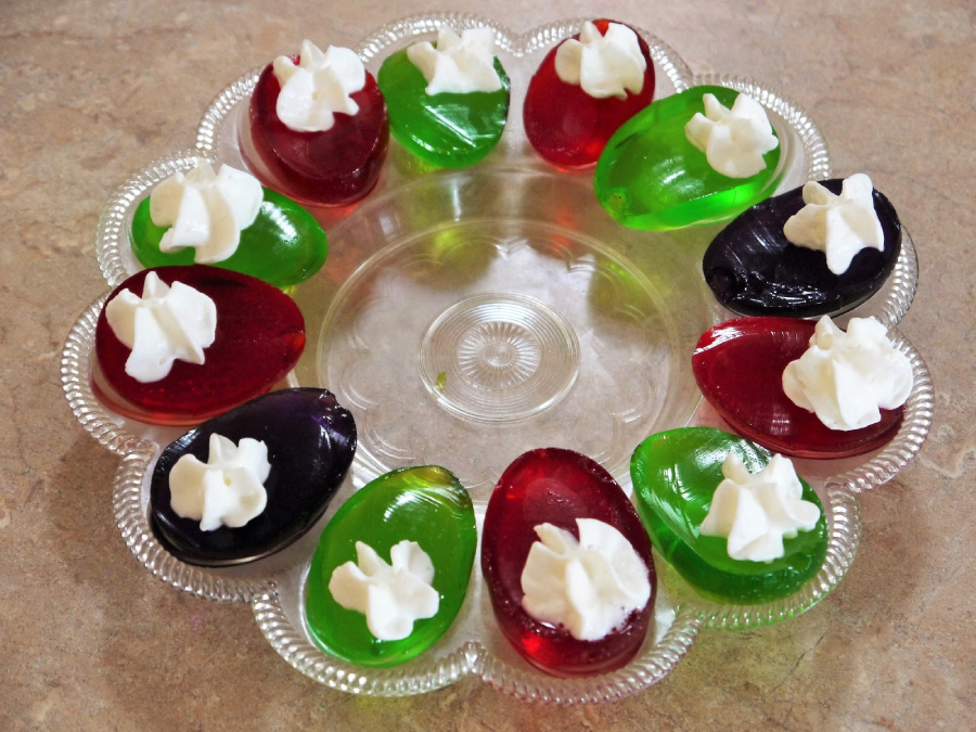 A glass plate with red, green, and purple jelly slices, each topped with a dollop of whipped cream, arranged in a circular pattern on a beige surface.