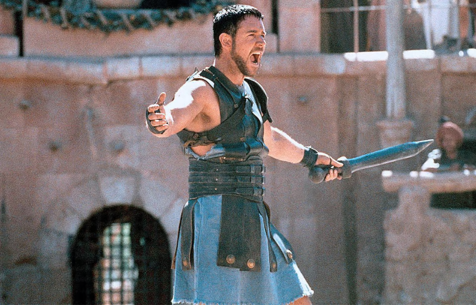 A man dressed as a Roman gladiator, wearing armor and a blue tunic, stands in an arena holding a sword and shouting with arms outstretched. The background shows stone walls and blurred spectators.