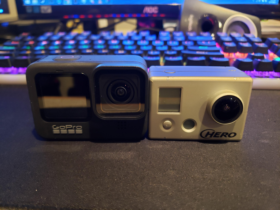 Two GoPro cameras, one modern black model and one older silver model, are placed side by side on a desk in front of a colorful mechanical keyboard and a computer monitor.