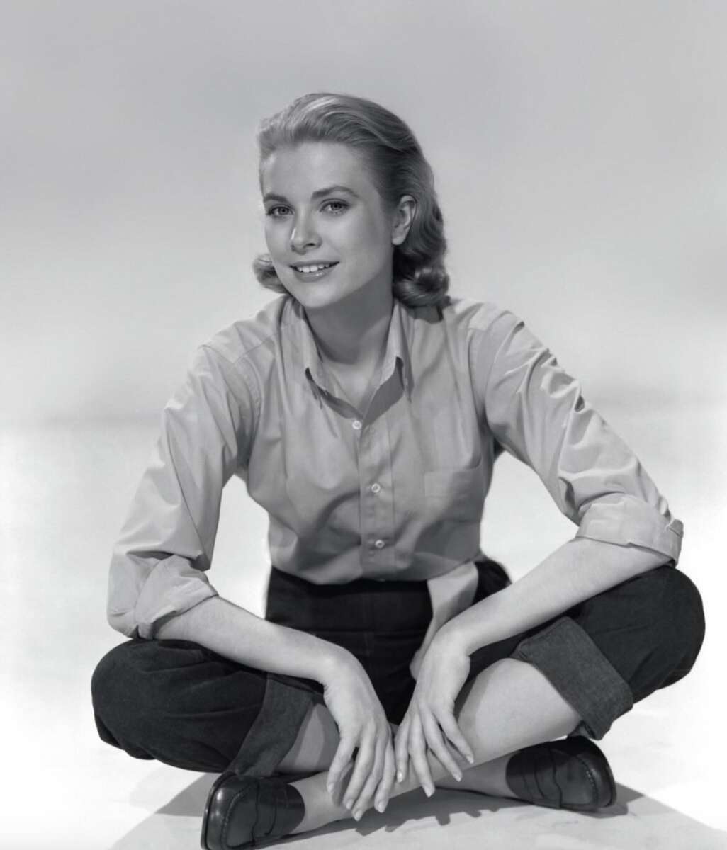 A woman with blonde, shoulder-length hair sits cross-legged on the floor, wearing a button-up shirt with rolled sleeves, dark pants, and loafers, smiling softly against a plain background.
