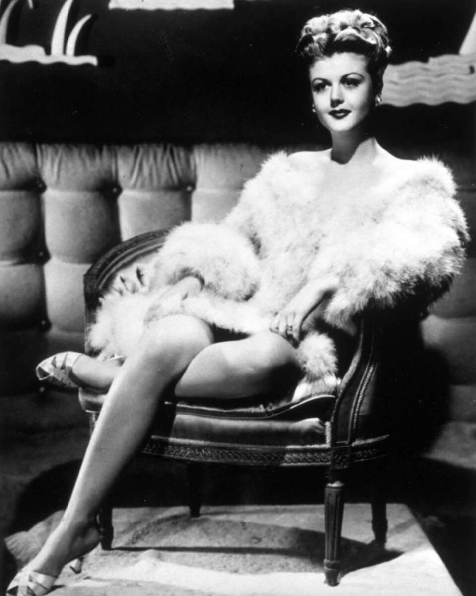 A glamorous woman in a fur-trimmed robe sits elegantly on a tufted chair, crossing her legs and smiling, with vintage decor in the background, in a black-and-white photograph.
