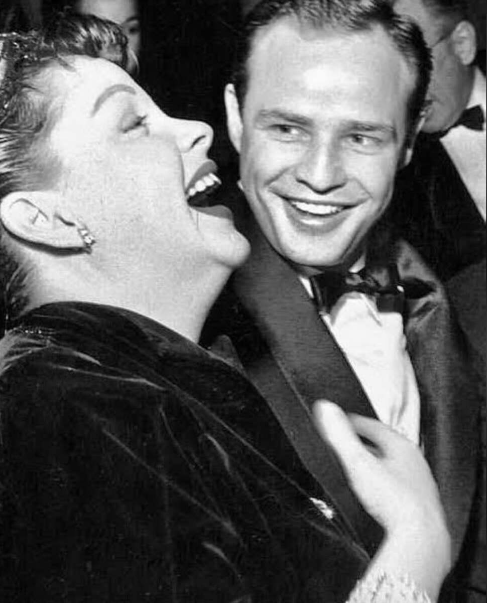 A woman laughing joyfully next to a smiling man in a tuxedo at what appears to be a formal event or party.