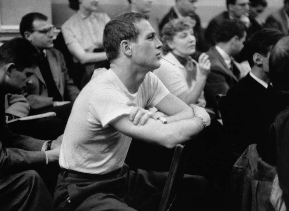 A man in a white t-shirt sits with his arms crossed, leaning forward and listening intently among a group of seated people in a crowded room. The setting appears formal, and most others are dressed in business attire.