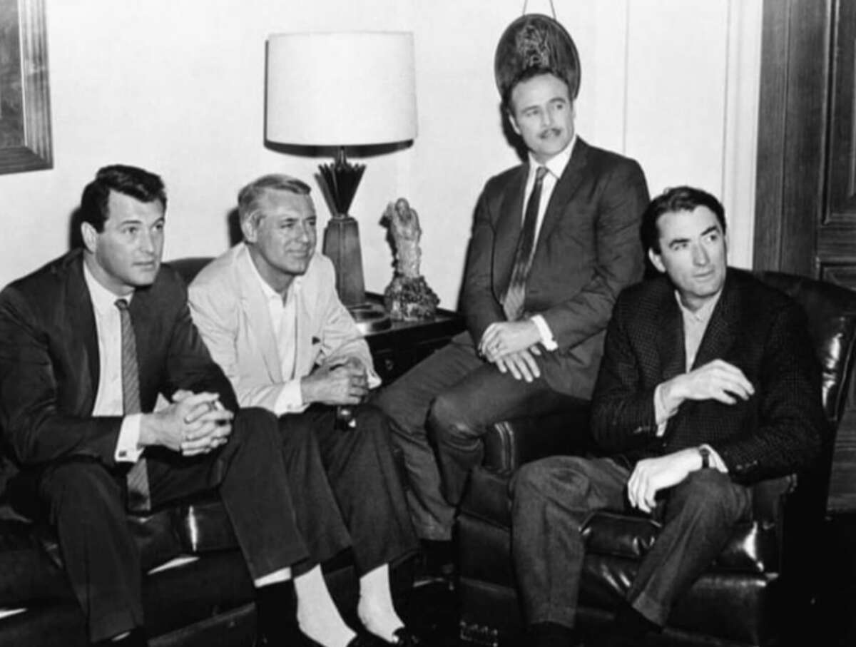 Four men in suits sit together in a living room, two on a sofa and two on armchairs, with a lamp and decorative items on a table behind them. The scene appears to be from the mid-20th century.
