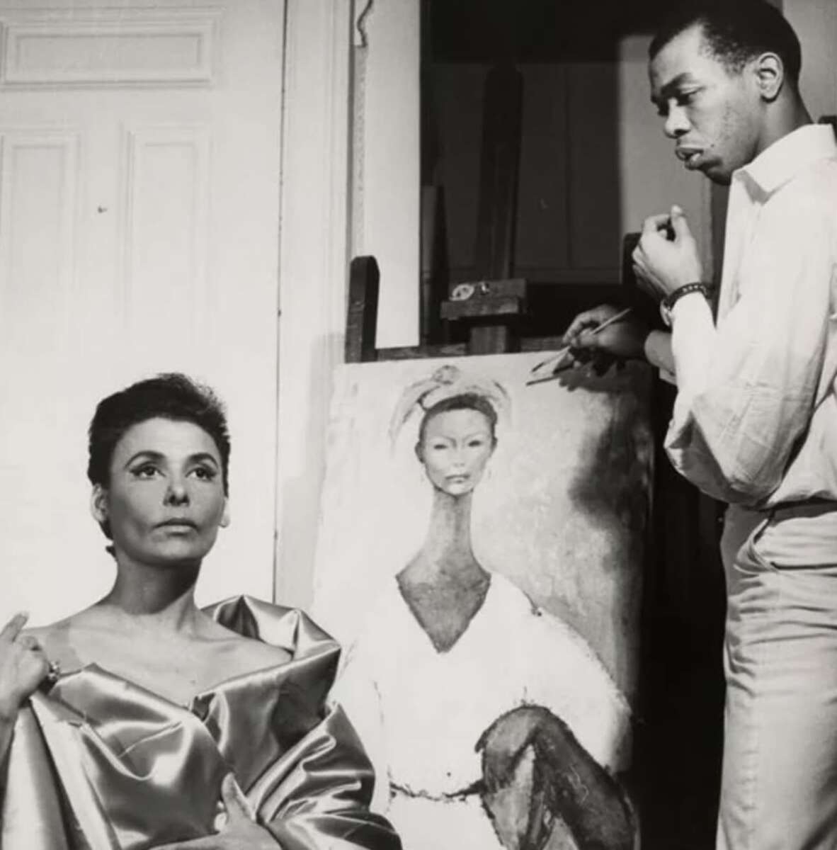 A woman draped in shiny fabric poses beside an artist painting her portrait. The artist stands thoughtfully with a brush, and the unfinished portrait on the easel resembles the woman’s pose and features.