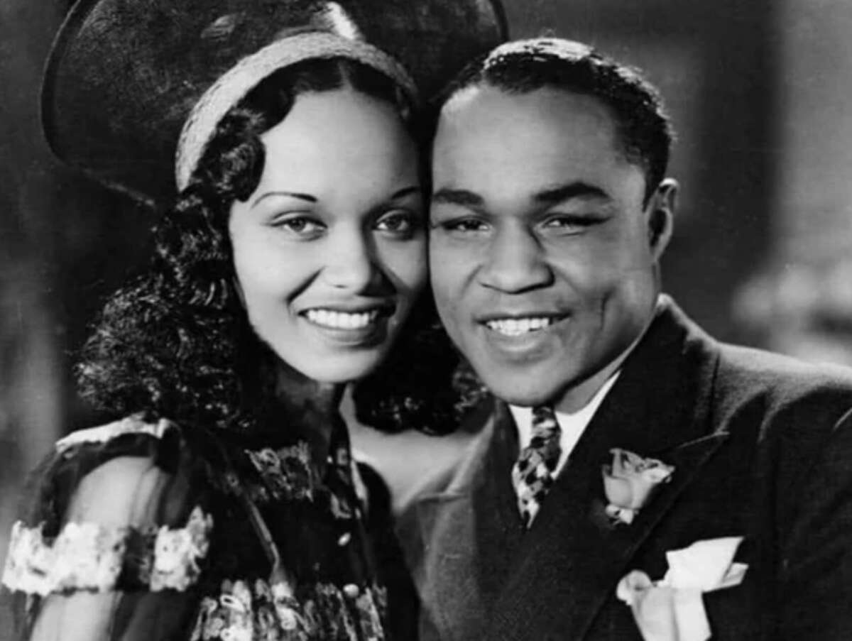 A smiling woman in a vintage dress and hat stands next to a smiling man in a suit and tie, both posing closely together for a black and white portrait.