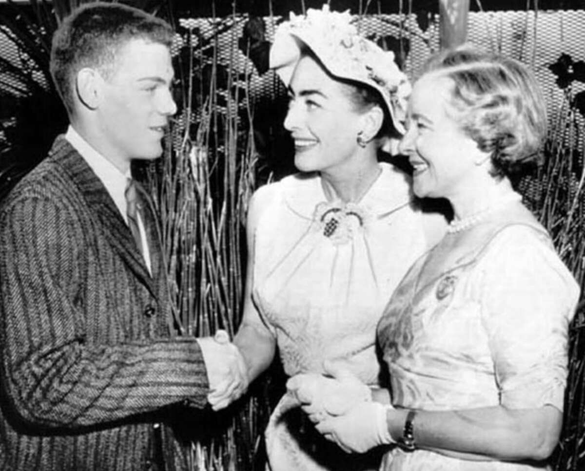 A young man in a suit shakes hands with a smiling woman in a hat and dress. Another woman in a light dress stands beside them, also smiling. The group appears to be at a formal event or gathering.